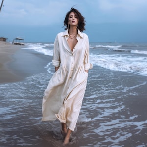 May include: A long, cream-coloured button-down dress with long sleeves and pockets. The dress is worn on a beach with the ocean in the background. The model is walking in the water.