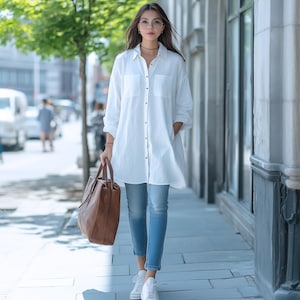 May include: A woman wearing a white oversized button-down shirt, blue jeans, and white sneakers. She is carrying a brown leather tote bag.
