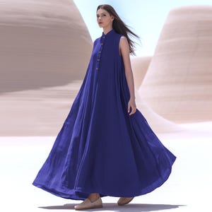 Pode incluir: Vestido azul longo sem mangas com gola mandarim e botões frontais. O vestido é feito de um tecido fluido e é usado com sapatos rasos bege. O fundo apresenta uma paisagem desértica com dunas de areia.