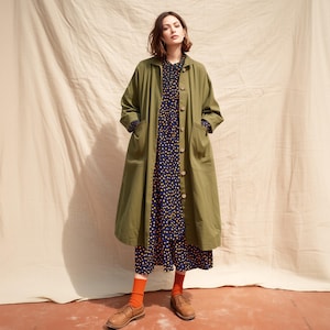 May include: A woman in an olive green long coat over a navy blue polka dot dress. The outfit is completed with orange socks and brown leather shoes. The coat has large pockets and buttons down the front.