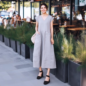 Könnte beinhalten: Eine Frau trägt ein hellgraues Leinenkleid mit Taschen und schwarzen Schuhen mit Absatz. Sie steht vor einem Café mit Pflanzen in Töpfen.