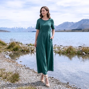 May include: A long, emerald green dress with short sleeves and a round neckline. The dress has pockets and is worn with beige heels. The background features a lake and mountains.