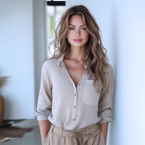 May include: A woman with long, wavy brown hair is wearing a light beige button-down shirt with a chest pocket and a pair of beige high-waisted pants.