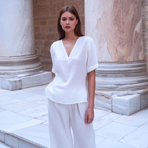 Könnte beinhalten: Eine Frau trägt ein weißes Oberteil mit V-Ausschnitt und kurzen Ärmeln sowie eine passende Hose mit weitem Bein. Das Outfit besteht aus leichtem Stoff. Der Hintergrund zeigt große weiße Säulen und einen Marmorboden.