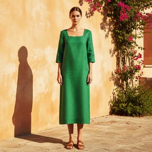 Peut inclure: Une femme porte une longue robe en lin vert émeraude avec un col carré et des manches trois-quarts. La robe arrive à mi-mollet. Elle porte des sandales marron. Le fond est un mur texturé orange clair avec une vigne fleurie.