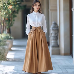 May include: A woman wearing a white button-down shirt and a long, flowing, tan skirt with a matching tie belt. The skirt has deep pockets. The outfit is worn outdoors in a light-filled, architectural setting.