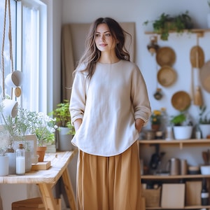May include: A woman wearing a white linen top with a relaxed fit and a brown skirt. She is standing in a room with a window and plants.