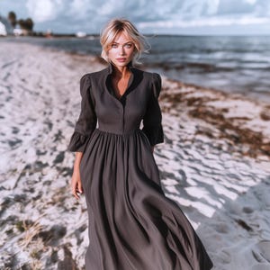 Könnte beinhalten: Ein dunkelgraues, geknöpftes Kleid mit hohem Kragen und langen Ärmeln. Das Kleid hat einen fließenden Rock und wird an einem Sandstrand mit dem Meer im Hintergrund getragen. Das Kleid ist aus leichtem Stoff gefertigt.