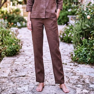 Può includere: Un paio di pantaloni marroni a tinta unita con taglio a gamba dritta e una camicia abbottonata coordinata con maniche a tre quarti. I capi sono realizzati in un tessuto testurizzato e includono tasche laterali, formando un set coerente.
