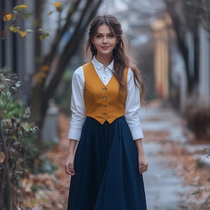 Può includere: Una donna indossa una camicia bianca con un gilet giallo senape e una gonna blu scuro. È in piedi su un sentiero con foglie cadute.