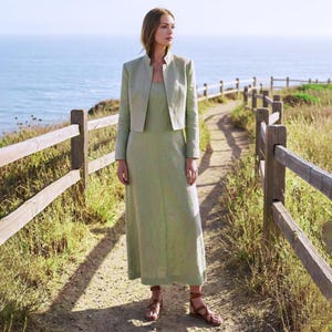 May include: A light green linen dress and matching cropped jacket. The dress is a simple, sleeveless design, and the jacket has a mandarin collar. The outfit is paired with brown leather sandals. The background features a wooden fence, a path, and the ocean.