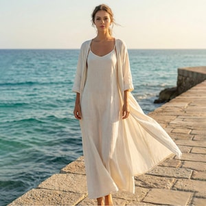 May include: A woman wearing a cream-colored linen dress and a matching long, open cardigan. The dress has spaghetti straps and a flowing silhouette. The outfit is worn on a stone pier with the ocean in the background.