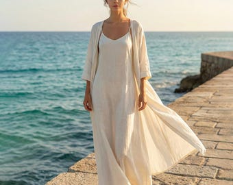 May include: A woman wearing a cream-colored linen dress and a matching long, open cardigan. The dress has spaghetti straps and a flowing silhouette. The outfit is worn on a stone pier with the ocean in the background.