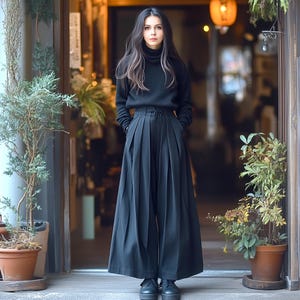 May include: A woman wearing a black turtleneck sweater and black wide-leg pants with pleats. She is standing in front of a doorway with plants on either side.