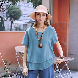 May include: A light blue linen top with a layered design and short sleeves. The top features a wooden elephant pendant necklace with brown and green beads.