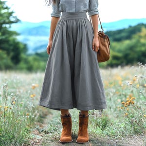 Puede incluir: Una falda de lino gris con cintura alta y pliegues. La falda es de largo hasta la rodilla y tiene una silueta fluida. La modelo lleva botas de tobillo marrones.