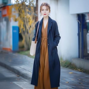May include: A long navy blue trench coat worn over a solid brown V-neck jumpsuit with a tie waist. The outfit is paired with a cream-colored tote bag featuring brown leather straps. The coat has a notched lapel and long sleeves with a button closure.