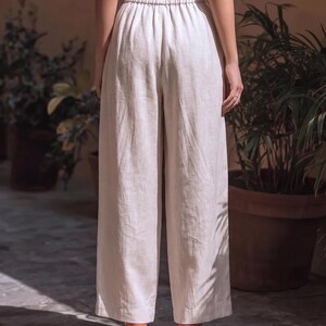 May include: Cream-colored linen pants with a wide leg and elastic waistband. The pants are paired with orange high heels. The image is taken from a rear view, showcasing the outfit's silhouette and texture.
