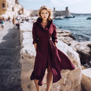 Puede incluir: Un vestido largo y fluido con capucha de color burdeos y mangas largas. El vestido está abierto por delante y ceñido a la cintura con un lazo. La modelo está de pie sobre un muro de piedra junto al mar.