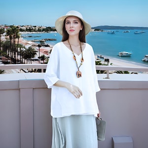 May include: A white linen top with a v-neck and three-quarter sleeves. The top is paired with a light gray linen skirt and a straw hat. The woman is wearing a necklace with brown and orange beads.