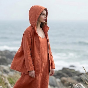 May include: A rust-colored hooded jacket and dress ensemble. The jacket has a button-up front and a drawstring hood. The dress has a V-neck and a flowing silhouette. The outfit is made of a lightweight fabric, perfect for layering. The background features a seascape.