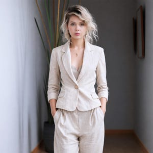 May include: A beige linen suit consisting of a blazer and matching pants. The blazer has a single button closure and pockets. The pants are high-waisted with a relaxed fit. The suit is worn by a person with blonde hair.