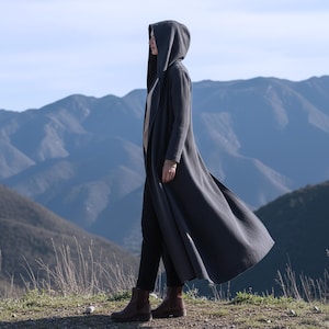 May include: A long, dark gray hooded coat with a flowing design. The coat is worn over a light-colored top and black pants. The person is standing on a grassy area with mountains in the background.