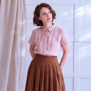 Peut inclure: Une chemise rose clair à manches courtes et un jupe plissée marron. La chemise a un devant boutonné et la jupe est de longueur midi. L'ensemble est fait d'un tissu doux et naturel.