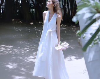 May include: A sleeveless white dress with a V-neck and a matching tie belt. The dress is floor-length and paired with white shoes. A small bouquet of pink and white flowers is held in the hand.