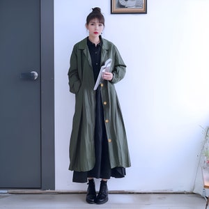 May include: A long, green, button-up coat with a black dress underneath. The coat has a collar and pockets. The person is wearing black boots.