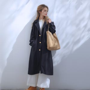 以下が含まれることがあります： 金色のボタンが付いた長い黒色のトレンチコート。コートは白いシャツと白いパンツの上に着用されています。その人は薄い茶色のトートバッグを持っています。