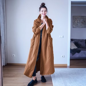 May include: A woman in a long, brown coat with large pockets. The coat is open, showing a black top and trousers. She wears black shoes and has her hair up. The coat looks to be made of linen or a similar fabric. The coat's length is approximately 140 cm.