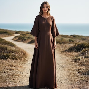 Könnte beinhalten: Ein langes, braunes Kleid mit weiten Ärmeln und rundem Ausschnitt. Die Person trägt das Kleid auf einem Feldweg, mit dem Meer im Hintergrund. Das Kleid scheint aus einem Naturstoff wie Leinen oder Baumwolle zu sein.