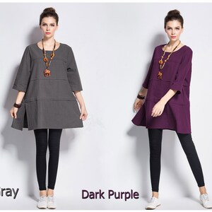 May include: Two women wearing loose-fitting, 3/4 sleeve shirts with a simple, round neckline. The shirt on the left is gray and the shirt on the right is dark purple. Both women are wearing black leggings and white sneakers.