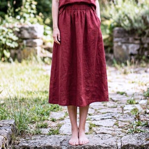 May include: A burgundy-colored linen skirt with an elastic waistband and pockets. The skirt falls to mid-calf length. The person is standing on a stone path in a garden setting.