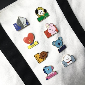 May include: A collection of eight enamel pins featuring cartoon characters Van, Chimmy, Cooky, Tata, Koya, Shooky, RJ, and Mang, each attached to a white canvas tote bag with black vertical stripes. The pins display colorful character designs.