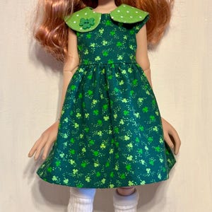 May include: A doll wearing a green dress with a clover pattern, a green bow, and white leg warmers. The doll has red hair, green eyes, and pink shoes. The dress has a white collar with green polka dots.