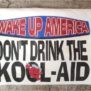 May include: White t-shirt with a red, white, and blue banner that says "WAKE UP AMERICA". Below the banner is black text that says "DON'T DRINK THE KOOL-AID" with a red Kool-Aid pitcher graphic.