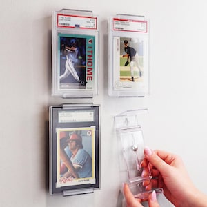 May include: Three clear acrylic wall-mounted display cases holding graded baseball cards, including a 1992 Fleer Jim Thome, a 2002 Topps Ten Randy Johnson, and a 1978 Topps Pete Rose card, alongside an empty clear acrylic display mount for wall installation.