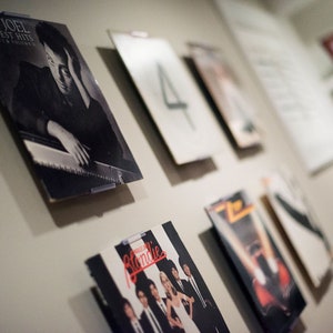 May include: A collection of vinyl record covers displayed on a wall, including Billy Joel's Greatest Hits Volume I & II, a cover with the number 4, and Blondie's Parallel Lines, featuring a black and white photograph of the band members.