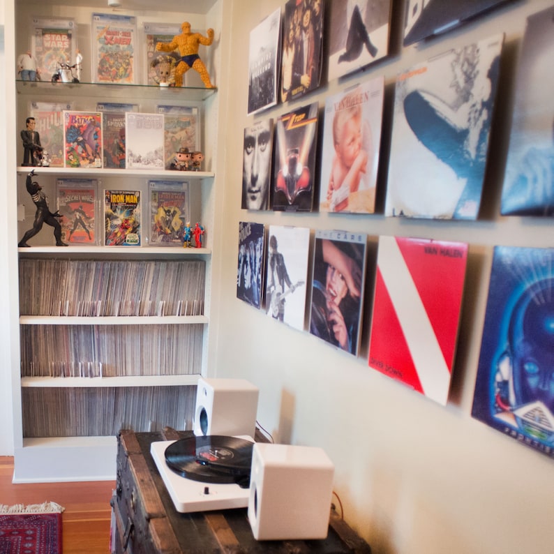 May include: A white record player with two white speakers sits on a dark wooden trunk. A tall white shelving unit displays numerous comic books in protective cases and various figurines. A wall features a collection of vinyl record album covers.