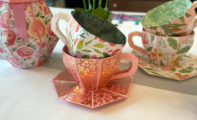 May include: A set of three pink and white teacups with floral patterns. The cups are stacked on top of each other and the bottom cup is on a matching saucer.