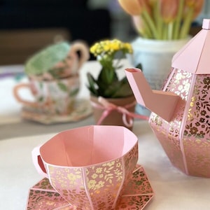 May include: A pink and gold tea set with a floral pattern. The teacup and saucer are made of paper and have a delicate design. The teapot is also made of paper and has a more intricate design.