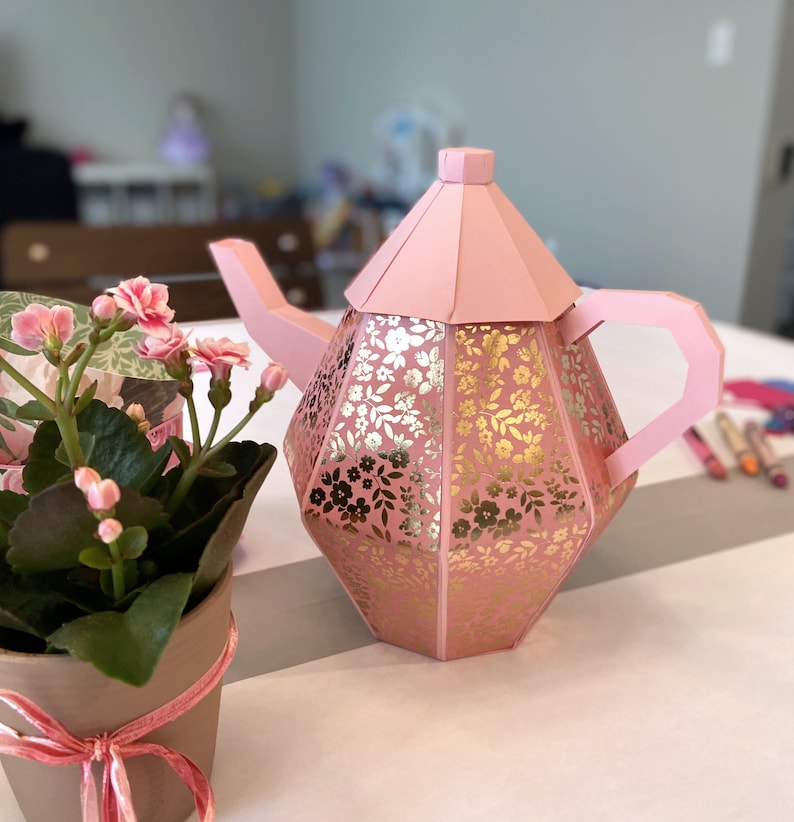 May include: A pink teapot with a gold floral pattern. The teapot has a lid and a handle. The teapot is sitting on a white surface.