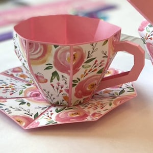 May include: A pink teacup and saucer with a floral pattern. The teacup has a handle and the saucer is round. The teacup and saucer are made of paper.
