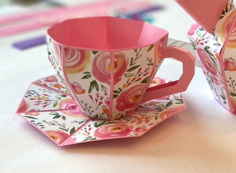 May include: A pink teacup and saucer with a floral pattern. The teacup has a handle and the saucer is round. The teacup and saucer are made of paper.