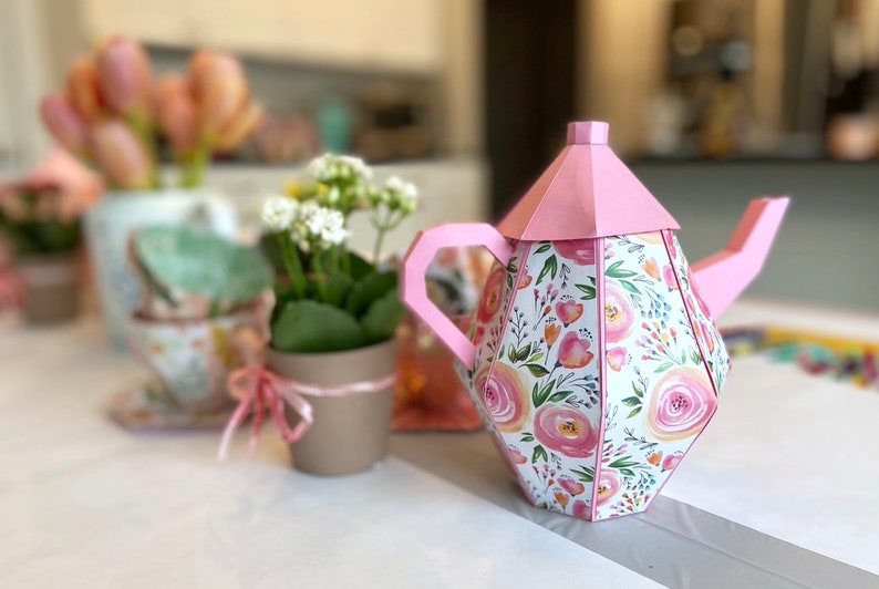 May include: A pink and white floral teapot with a pink lid. The teapot is made of paper and has a geometric design.