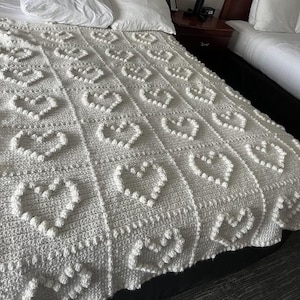 May include: A white crocheted blanket featuring a repeating grid pattern of raised heart shapes. The blanket is constructed from individual square panels, each displaying a textured heart design created with bobble stitches on a flat surface.