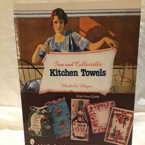 以下が含まれることがあります： ミシェル・ヘイズ著「Fun and Collectible Kitchen Towels」というタイトルの本。表紙には、タオルを持った女性のヴィンテージイラストと、その下にキッチンのタオルの例が描かれています。この本には価格ガイドが含まれています。