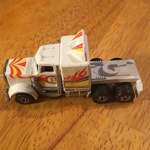 May include: White toy truck with red and yellow flames and a Simpson logo. The truck has a gray flatbed and black wheels.